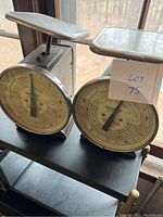 Front view of two antique postal scales side by side showing round dial faces, metal trays, and some surface wear.