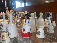 Photo of 12 porcelain and glass decorative bells arranged on glass shelf with reflection showing variety of figures and detailed paintwork.