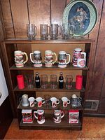 Full view of wooden bookshelf with Budweiser steins, glass mugs, Coca-Cola bottles, small coffee mugs and display containers arranged on three shelves.