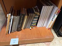Photo shows 16 cookbooks of various sizes and colors arranged upright on a wooden shelf. Titles include Secrets of Better Cooking, arts and crafts American cooking, and health-focused cookbooks.
