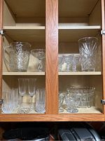 Photo showing upper and lower shelves of wooden cabinet displaying various pieces of crystal glassware including pitcher, vases, bowls, and stemmed glasses arranged inside.