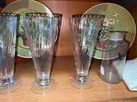 Four clear glass tumblers in front of two decorative plates and a glass jar with metal lid on a wooden shelf
