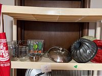 Shelf photo showing clear glass jars, stainless steel bowls, metal pot with lid, baking molds including bundt pan, red mold, kitchen utensils, and apron on left.