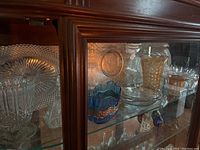 Photo showing various glass bowls and glassware in a wooden display cabinet. Includes carnival glass, blue glass, amber glass, and clear glass pieces with detailed patterns and shapes.