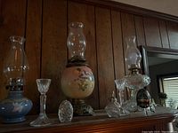 Wide view showing three hurricane lamps, several glass candle holders, crystal egg, Fabergé-like egg, and perfume decanter on wooden shelf