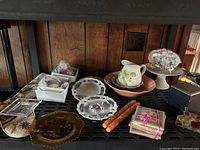 Wide view showing entire collection on a black metal shelf with wood panel wall background. Includes sushi plates, embroidered plates, decorative bowls and candles.
