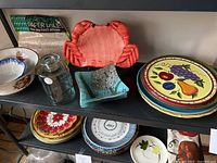 Multiple bowls, plates, glass jar, paper doilies displayed on shelves including red crab plate and fruit motif plates