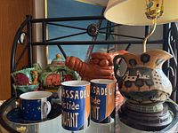 Photo showing pottery pitcher lamp, two Sango mugs, blue and gold cup with saucer, green pottery cup, and reddish-brown pottery piece.