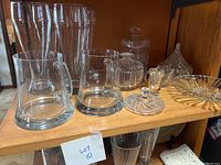 Front view of tall beer glasses, short handled glasses, lidded candy containers, etched glass candy dish, and shot glass on a wooden shelf