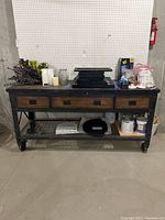 Front view of the full console work bench showing top wooden surface with multiple items placed on it, three wooden drawers, and metal wire mesh shelf underneath holding cans and other objects. Bench rests on four caster wheels.