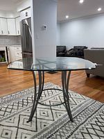 View showing glass tabletop and metal base with curved legs on a patterned rug in a living room with wood flooring.