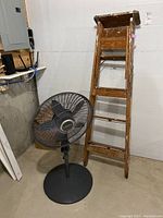 Wooden ladder standing next to black Lasko pedestal fan, showing front view of both items in basement setting.