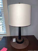Full view of the lamp showing the black metal base, bronze accent ring, and white fabric drum shade on a dark wooden table.