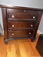 Front view of the wooden nightstand showing three drawers with metal knobs and carved corners at the base.