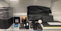 Overview of lot items arranged on shelving: Kenneth Cole black leather bag, black notebook, 3-hole punch, architectural tools, tape dispenser, bookends, calculator, wastebasket.