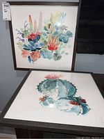 Photo showing two framed square floral-themed artworks, one with succulents and one with a flower bouquet on white background, placed beside a TV.