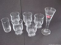 Front angled view of all 7 glasses including five with decorative bases, one plain glass and one tall Malteserkreuz branded glass.