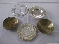 Five assorted bowls and stands displayed on a white surface, showing a mix of silver, silver-plated, brass, and glass materials.