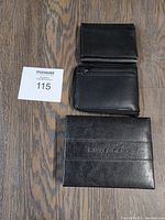 Three black leather men's wallets arranged on wood flooring, one wallet in Wenger branded box