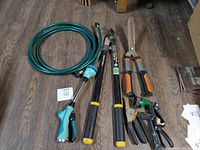 Image of garden tool lot including coiled green garden hose with spray nozzle, black and yellow loppers, orange and gray hedge shears, and two black and green pruning shears.