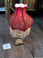 Photo showing full table lamp with red floral fabric shade and white tassels on brass metal base, with power cord and inline switch visible.