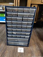 Front view of dark blue parts storage cabinet with multiple transparent plastic drawers on wood floor.