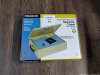 Box packaging showing Steelmaster fire-retardant security chest with lock and key inside, highlighting fire resistance up to 750°F.