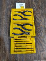 Top view of the open yellow hard plastic case showing 3 pairs of blue-handled pliers in the top two compartments and 8 precision screwdrivers in the bottom compartment.