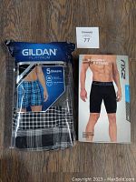 Packaged set of 5 Gildan Platinum boxers and 1 pair of packaged 2XU Accelerate compression shorts on wooden floor.