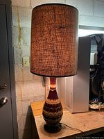 Full view of the vintage mid-century ceramic table lamp with original dark textured canvas shade, lit and placed on wooden surface against concrete wall.