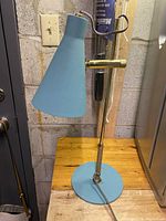 Blue shaded adjustable desk lamp with slim brass body and round base shown from side angle.