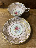 Porcelain cup and saucer showing gold leaf decoration, floral center, pink background, and scalloped edges.