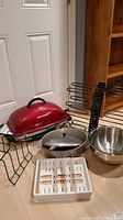 All items in the lot displayed together: red George Foreman grill, stainless steel roasting pot with lid, multiple black metal organizers for pots, pans and lids, white plastic organizer tray on wooden floor.