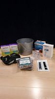 Photo showing the full lot items including plastic bucket, wrist braces, body sponges, Fitbit straps, hot and cold pad, shower curtain rings, and thermometer box