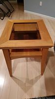 Front-facing photo showing light wood side table with beveled glass inlay and lower shelf on light hardwood floor.