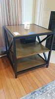 Side view of square side table showing wooden top and two shelves with black metal frame and crossed side bars.