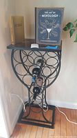 Wine rack with circular metal design, holding several bottles and two books on top, placed in a corner near a wall outlet.