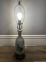 Front view of the vintage ceramic table lamp illuminated, showing floral painted design and metal harp for shade.
