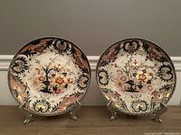 Pair of antique Crown Derby Imari plates displayed on metal stands, showing detailed floral designs in orange, black, and gold.
