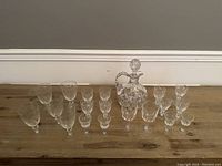 Wide photo showing the full set of 15 items: one decanter with stopper, 14 glasses of different sizes and designs on wooden surface.