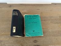 Two Hebrew books side by side on a wooden surface. One is a larger, hardcover dictionary with Hebrew text on the spine and cover. The other is a smaller green paperback with a visibly worn cover.