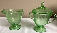 Creamer and lidded sugar bowl set made of uranium green Hazel Atlas depression glass with vintage Royal Lace pattern, shown side-by-side from front angle.