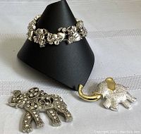 Elephant bracelet displayed on black stand showing multiple linked elephant charms, elephant brooch featuring circular bead-like decorations, and an elephant pendant with textured surface and gold-toned tusk.