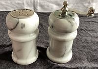 Front view of white marble salt shaker with metal perforated top and pepper grinder with metal grinding handle, both approximately 4 inches tall and 2 inches wide.