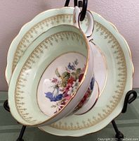 Wide mouth teacup hanging on a stand with matching saucer. Light green rim with gold detail and floral center pattern.