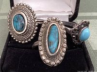 Lot of three turquoise rings displayed in a black ring holder with white cushion, showing different stone shapes and silver metal decorations