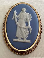 Front view of the vintage Wedgwood brooch showing the oval blue jasperware with white relief of a standing woman holding a garland, bordered by a twisted gold-tone frame.