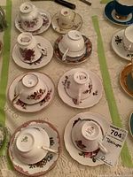 Photo showing eight bone china tea cups and saucers arranged on lace tablecloth, displaying floral patterns and gold trim, including brand marks on the undersides.