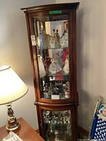 Front view of the tall dark wood display cabinet showing curved glass front door with brass hardware, glass shelves, and interior light illuminating porcelain figurines inside.
