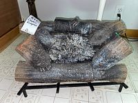 Full view of artificial electric fireplace logs set on black metal grate against white tiled floor and beige baseboard. Logs are dark brown with realistic wood texture and some white ash effect on the center logs.